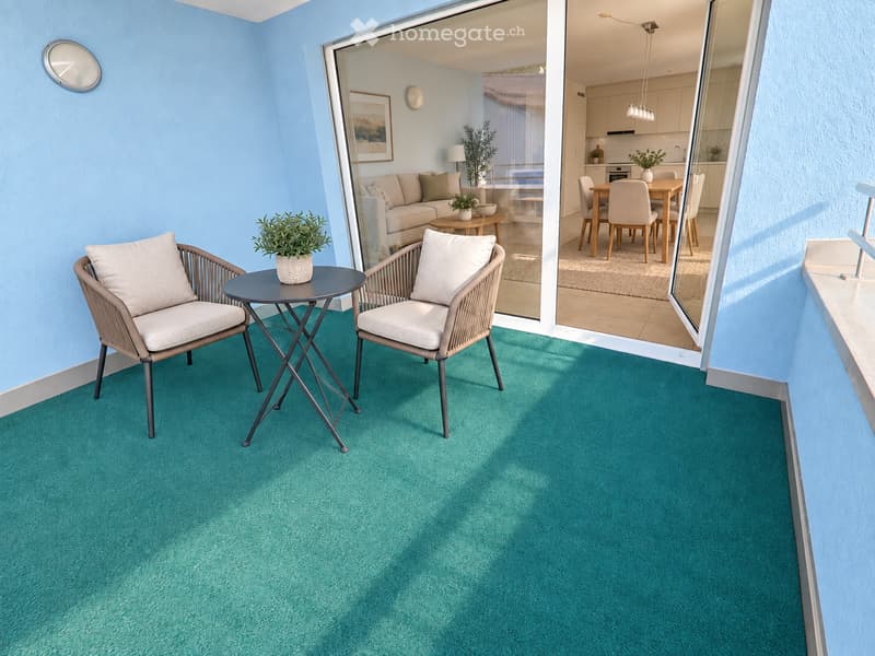 Balcony with two wicker chairs, a small round table, and a potted plant. The balcony has a turquoise carpeted floor and overlooks an interior space with a sofa, dining table, and kitchen area.
