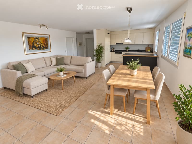 Open-plan living and dining area with a light grey sectional sofa, coffee table, dining table with chairs, and a modern kitchen area with cabinets and a counter. The space features tiled flooring, large windows, and decorative plants.