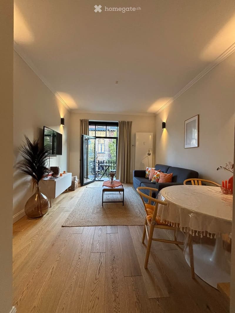 Bright living room with wooden flooring, a sofa, coffee table, dining table, and a TV. Large sliding doors open to a balcony.