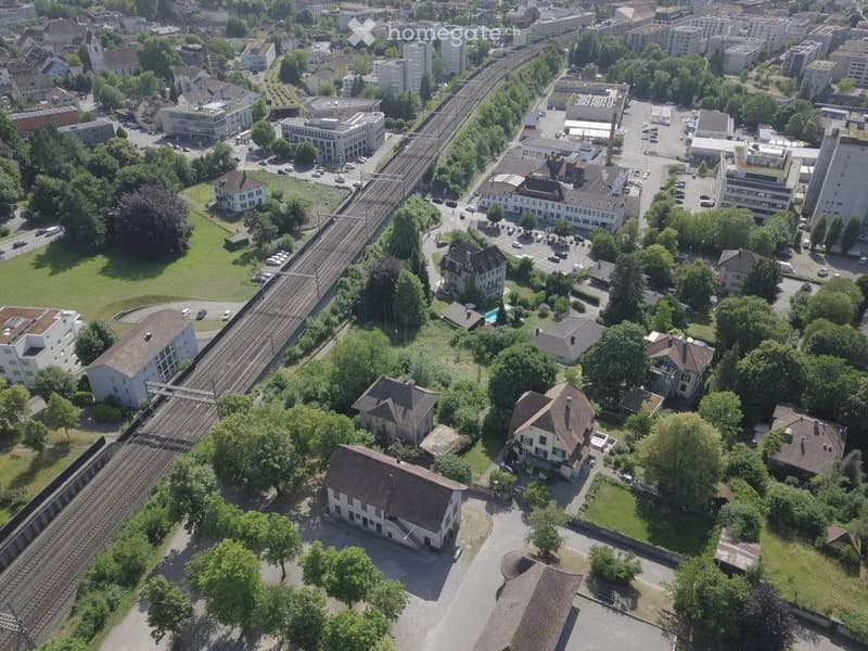 Aerial view of an urban area with multiple buildings, railway tracks, and green spaces.