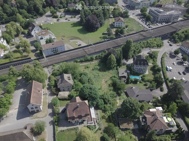 Aerial view of a residential area with multiple houses, a swimming pool, parking lots, and a railway line running through the neighborhood.