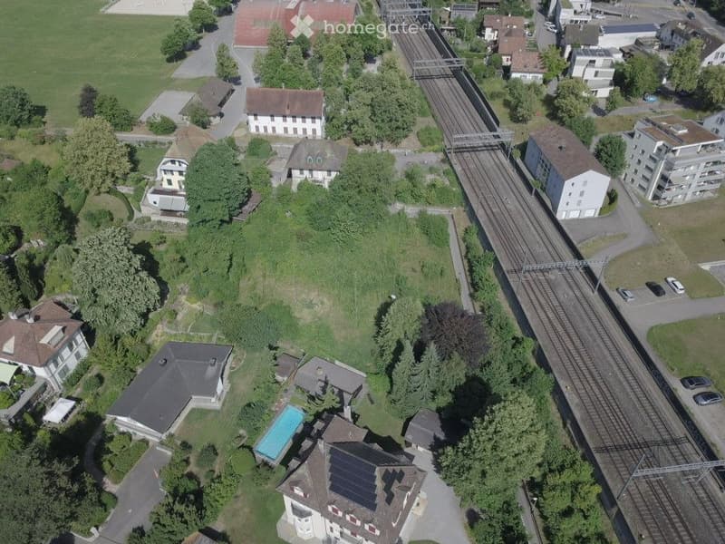 Aerial view of a residential area with multiple houses, a swimming pool, and a railway track running through the neighborhood.