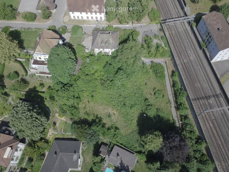 Aerial view of a residential area with multiple houses, gardens, a swimming pool, and a railway track running through the neighborhood.
