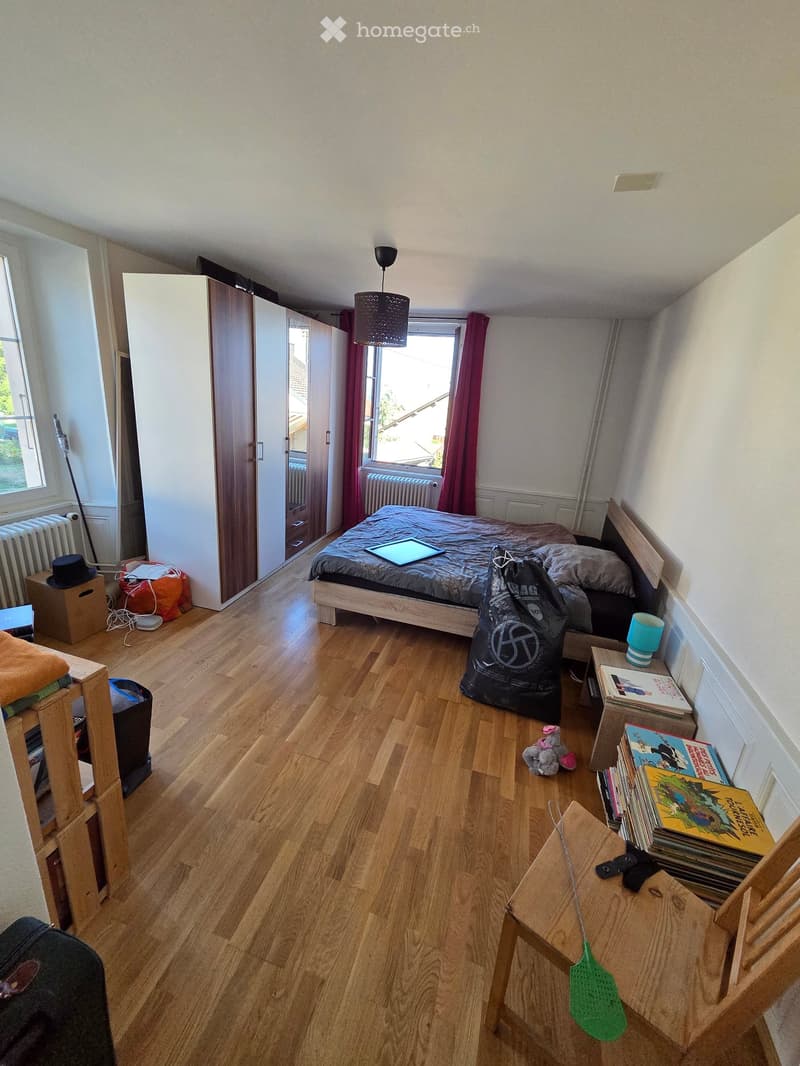 A small bedroom with wooden flooring, a bed with a dark duvet, a small table with books and a cup, a chair, a stool, a toy, and a table lamp.