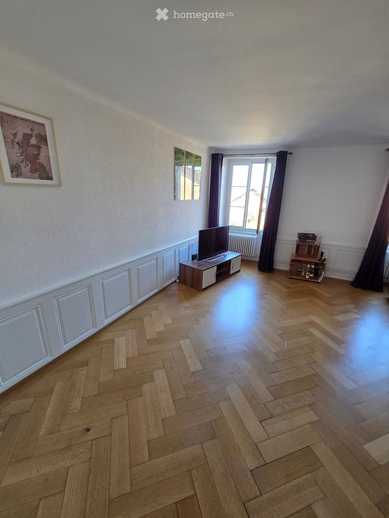 Living room with herringbone wooden flooring, large window with dark curtains, TV on a stand, small shelf with items, and framed map on the wall.
