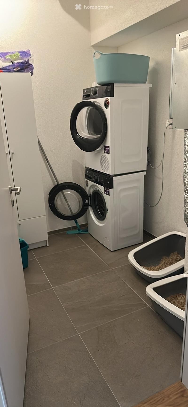 A small bathroom with tiled floor, white washing machine and dryer stacked, and two cat litter boxes.