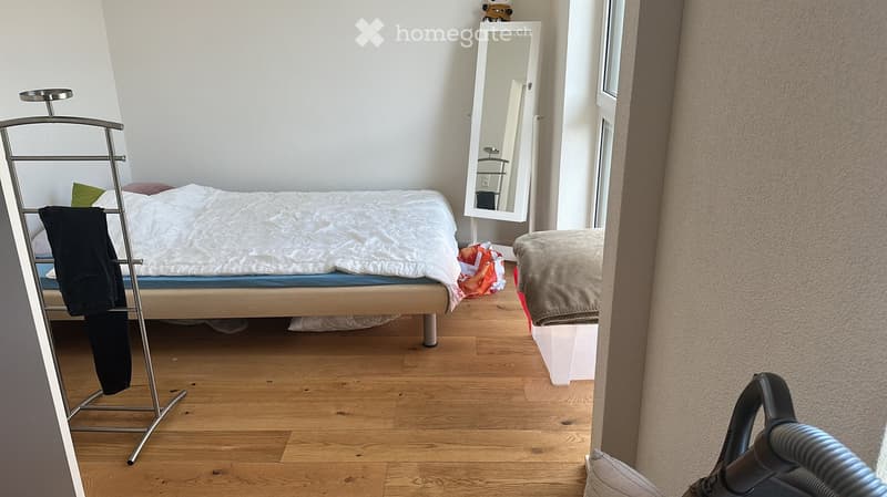 A small, neatly arranged bedroom with a single bed, a mirror, a clothing rack, and a storage box. The room features wooden flooring and white walls.