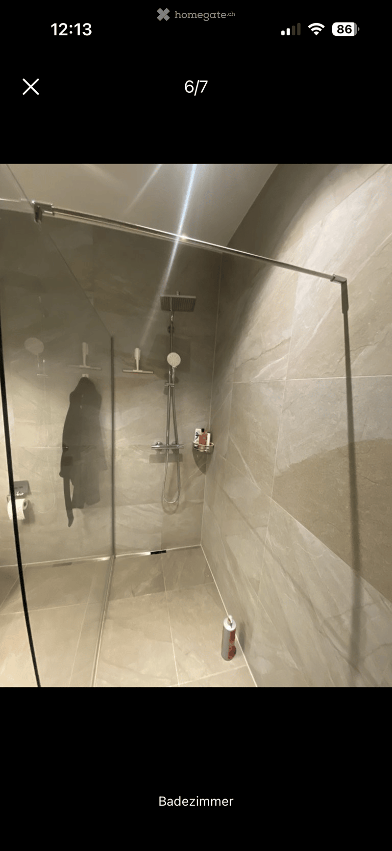 Modern shower area with glass enclosure, marble tiles, and a handheld showerhead. Includes a towel hanging on the glass door and a bottle of shower gel on the floor.