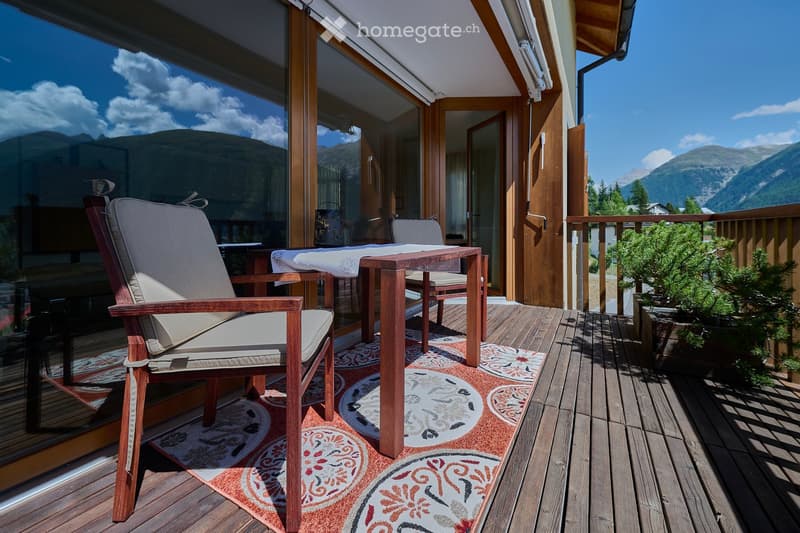 Balcony with wooden decking, two wooden chairs, a small table, and a patterned rug. Offers a scenic mountain view.