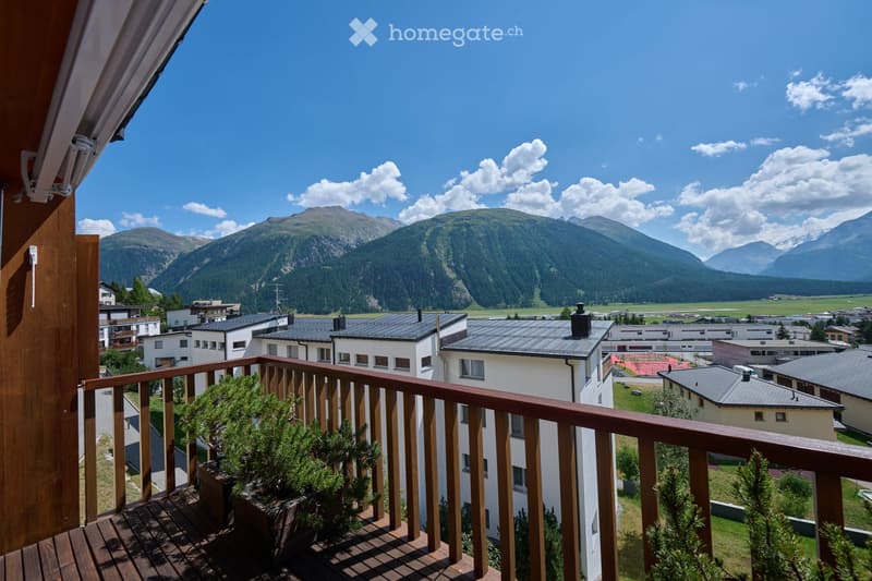 Balcony with wooden decking and railing, overlooking a town with buildings and distant mountains.