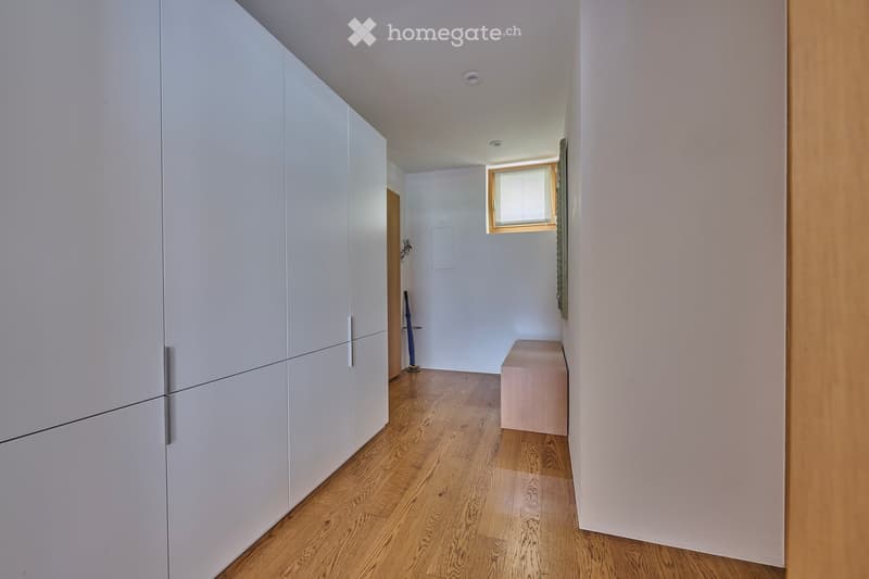 A narrow hallway with light wooden flooring, white walls, and built-in white storage units on one side. A small window is visible at the far end, and a small bench sits against the wall.