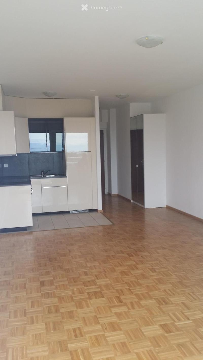Empty room with wooden flooring and white walls, featuring a modern kitchen area with white cabinets and stainless steel appliances.