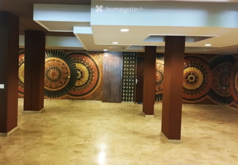 Spacious hallway with beige tiled flooring, wooden pillars, and decorative wall art featuring circular patterns in warm tones. The ceiling has recessed lighting, and there are doors visible along the walls.
