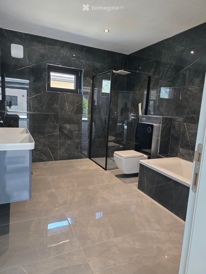 Modern bathroom with black marble walls and floors, featuring a large bathtub, toilet, sink, and glass-enclosed shower area.
