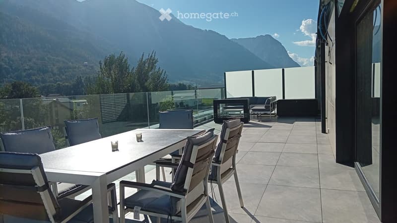 Modern outdoor terrace with a panoramic mountain view, featuring a white dining table with chairs, a sofa, and a glass railing.