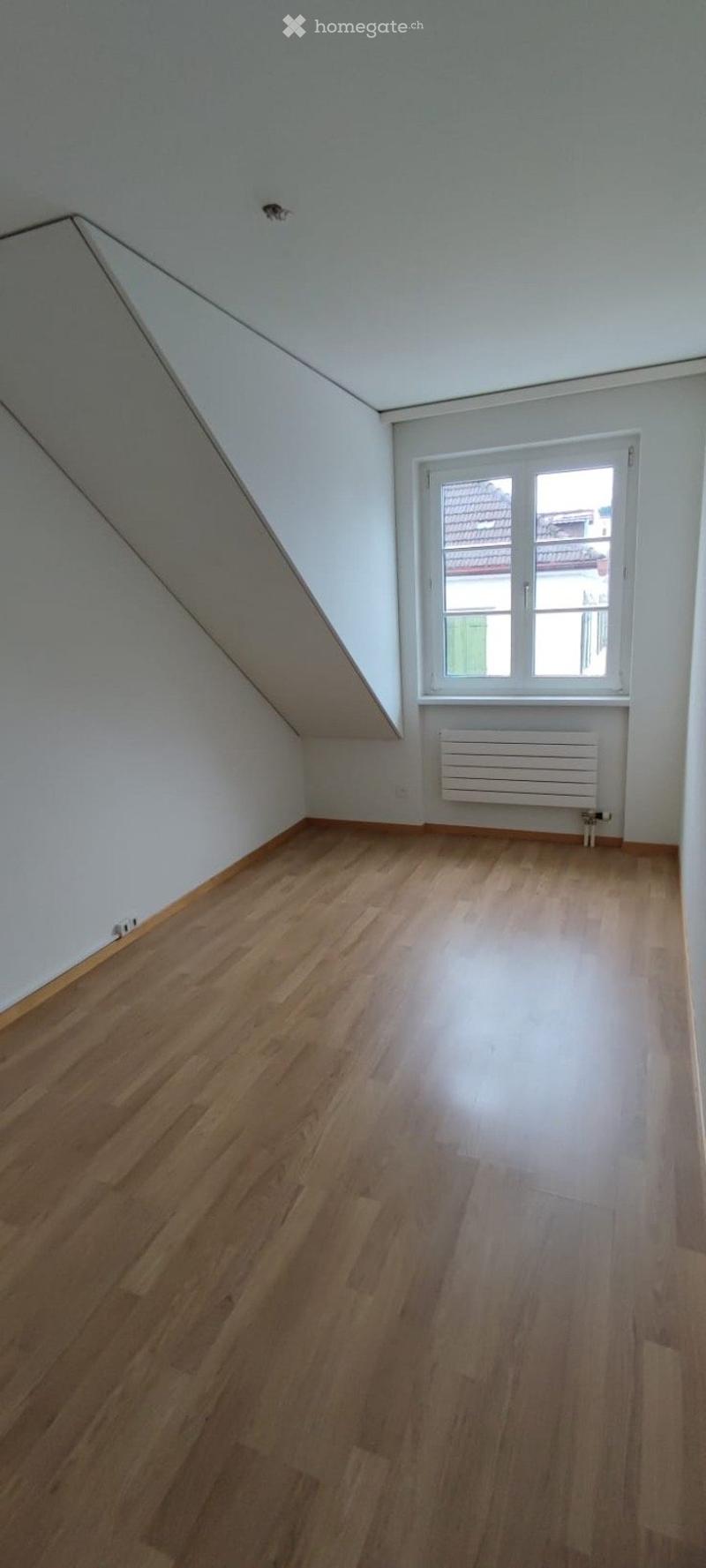Empty room with wooden flooring, white walls, and a window with a radiator below it. The ceiling has a slanted design typical of an attic space.
