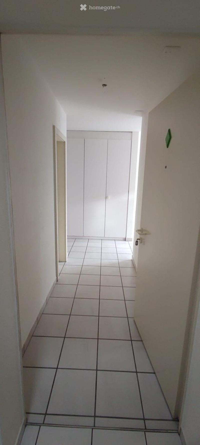 Empty hallway with white tiled floor and walls, featuring a closed double door at the end and a small shelf on the right side.