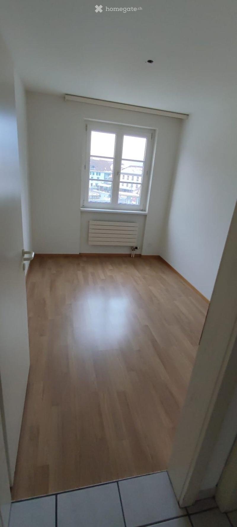 Empty room with wooden flooring, white walls, a window with a view, and a radiator below the window.