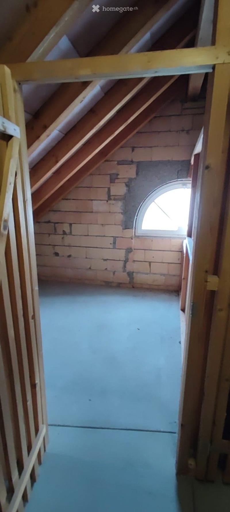 Empty attic space with exposed wooden beams, brick walls, and a small arched window letting in natural light.