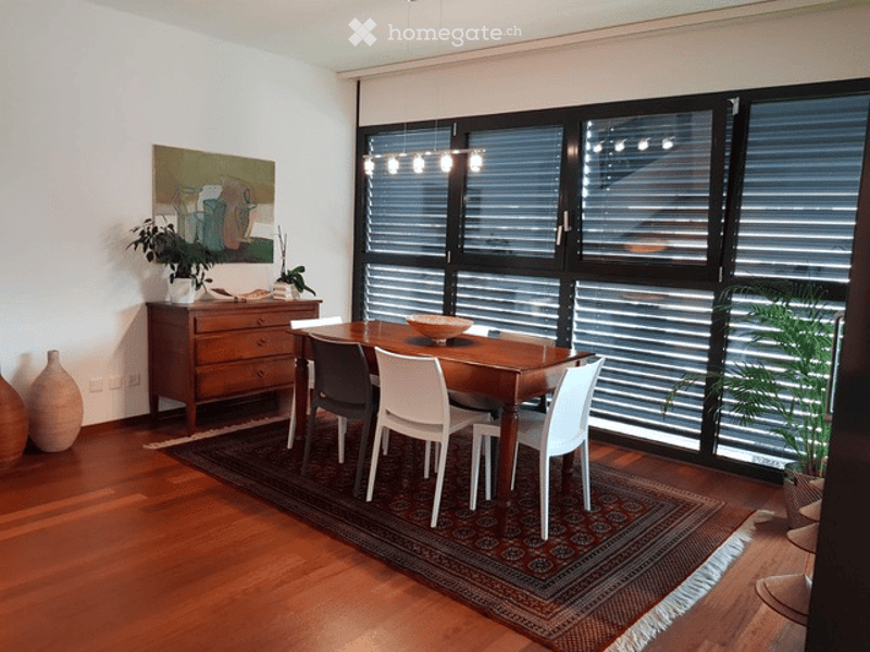 Dining room with wooden floor, large windows, dining table with chairs, cabinet, vases, and artwork.