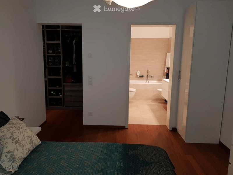 A bedroom with a bed, pillows, and a wardrobe. An adjacent bathroom is visible with a toilet, bathtub, and sink.