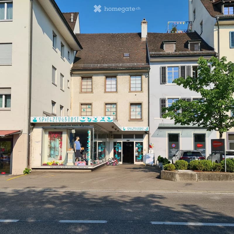 A two-story building with a storefront labeled "Spielzeugborse" and a sign reading "secondhand für klein & gross." The storefront displays toys and a mannequin, with a small bicycle and potted plants visible outside. The building has multiple windows and