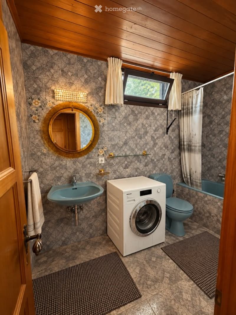 Bathroom with a blue sink, round mirror, washing machine, toilet, bathtub, and a window with curtains. Wooden ceiling and patterned tiles.