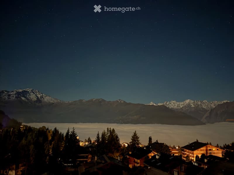 Nighttime view of a village nestled in a valley with snow-capped mountains in the background, illuminated houses, and a starry sky.