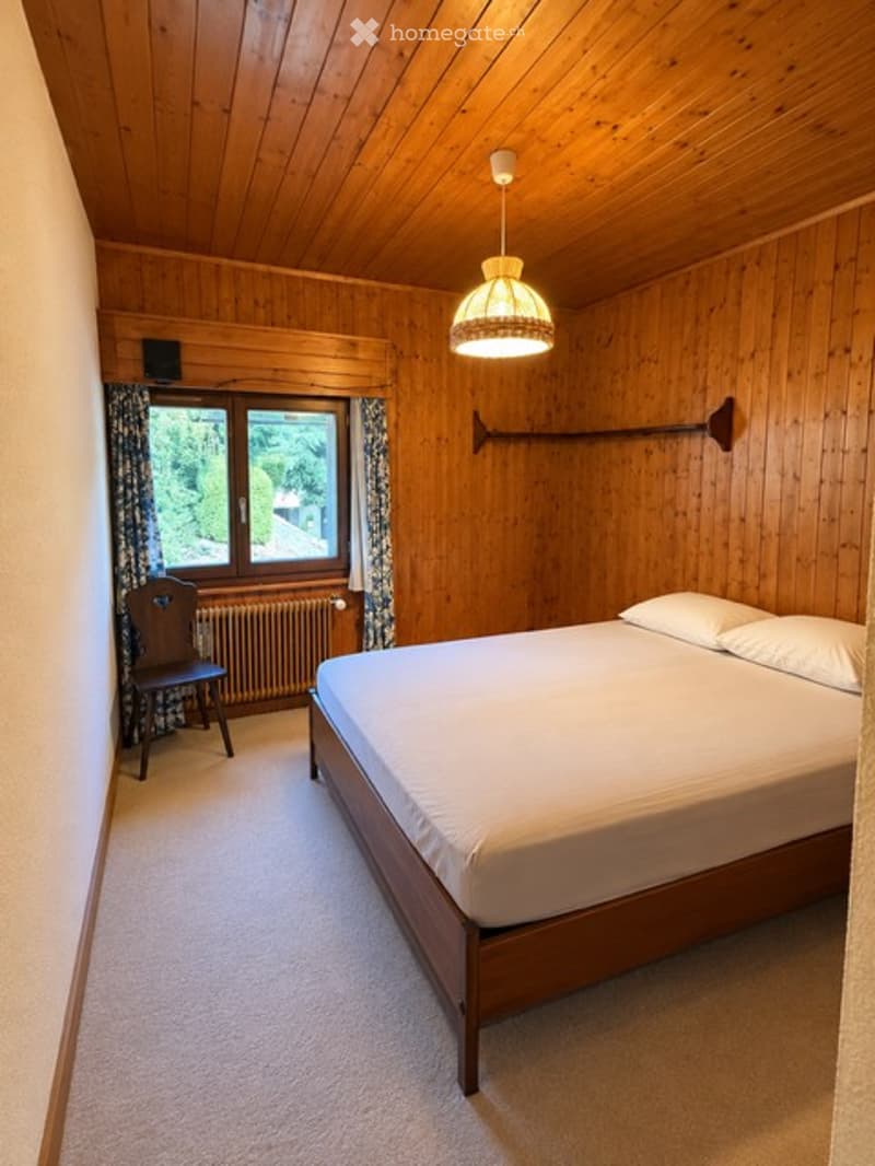 Cozy wooden-paneled bedroom with a double bed, wooden chair, radiator, and window with curtains.