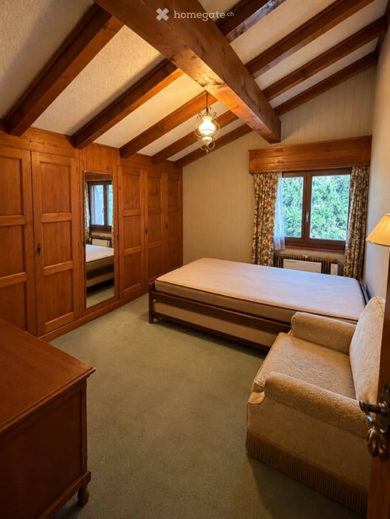 Cozy attic bedroom with wooden beams, a large bed, a sofa, a wardrobe, and a mirror. The room features exposed wooden beams on the ceiling and warm wooden paneling on the walls.