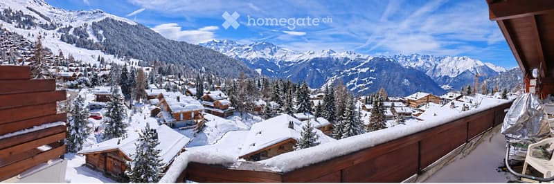 Balcony overlooking a snow-covered village with wooden houses and snow-capped mountains in the background.