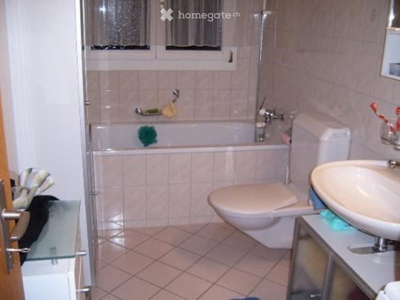 A small bathroom with a bathtub, toilet, sink, and tiled walls and floor. The bathtub has a showerhead above it, and there are small windows above the tub. A mirror is mounted on the wall above the sink.
