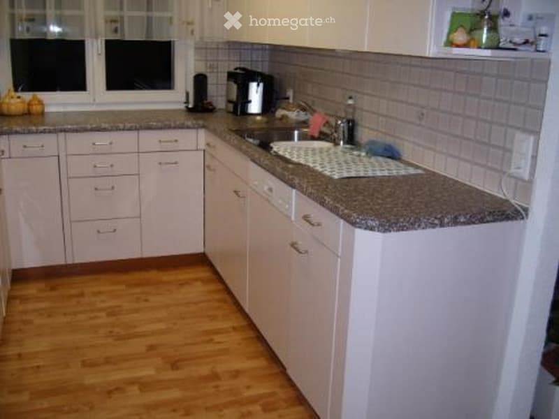 A modern kitchen with light-colored cabinets, granite countertop, and wooden flooring. Features include a sink, coffee maker, and various small appliances.