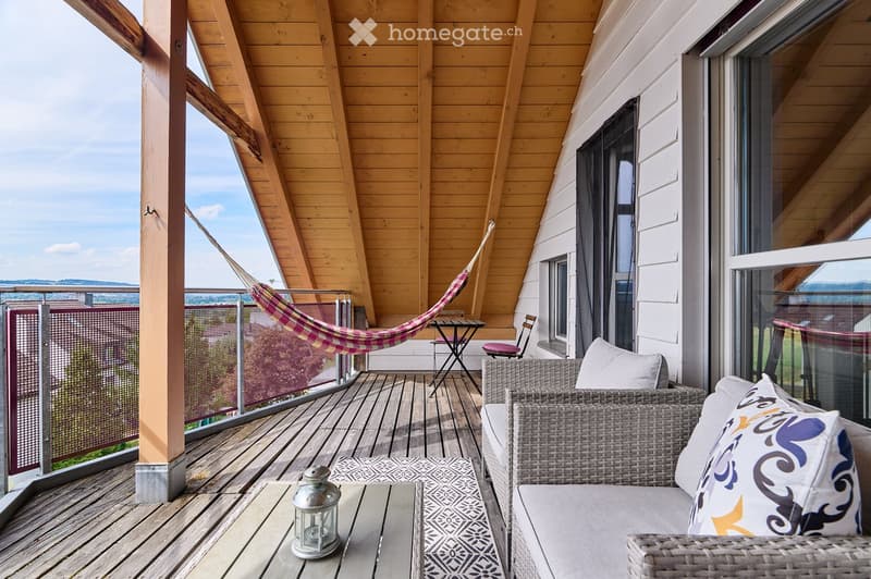 Wooden balcony with a view, featuring a wicker sofa, a small table, a chair, a hammock, a patterned rug, and a lantern.
