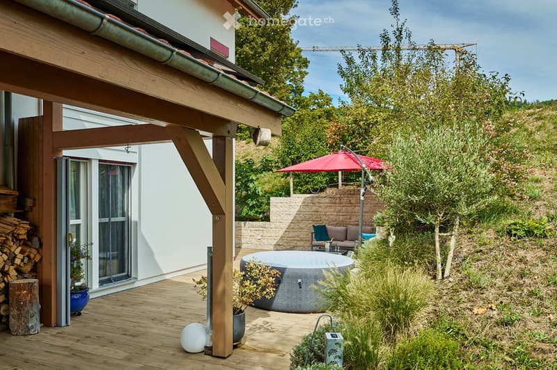 Outdoor garden area with a wooden deck, hot tub, seating area, umbrella, and various plants and trees.