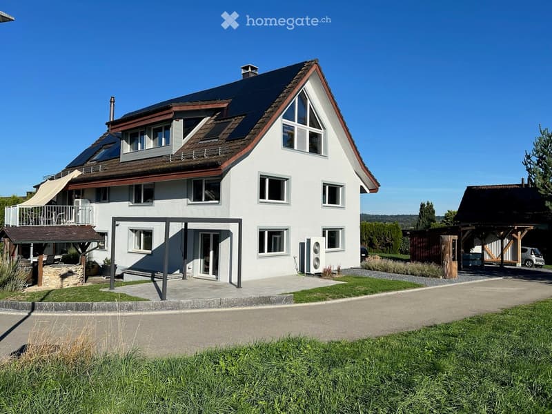 Two-story modern house with white exterior, solar panels on the roof, multiple windows, and a covered entrance area. There is a small patio with outdoor furniture and a stone fire pit.