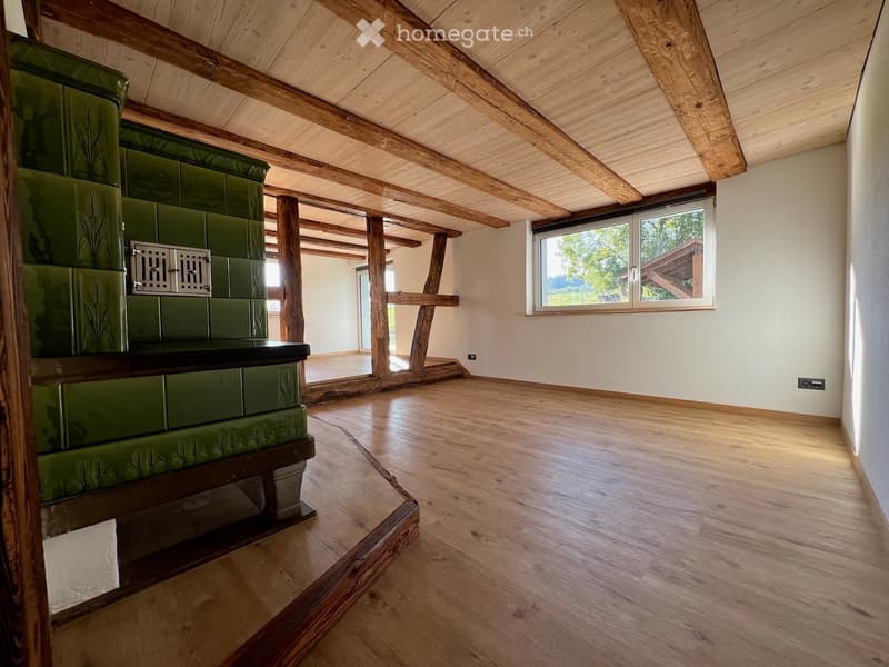 Empty room with wooden floor, exposed wooden beams, and a green tiled stove. Large window with view of greenery.