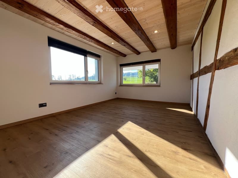 Empty room with wooden floor, white walls, exposed wooden beams on the ceiling, and two windows with blinds.