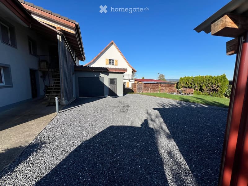 A two-story house with a garage, a small annex building, and a gravel driveway. The annex has a triangular roof and a small balcony. There is a wooden fence and a grassy area with benches in the background.