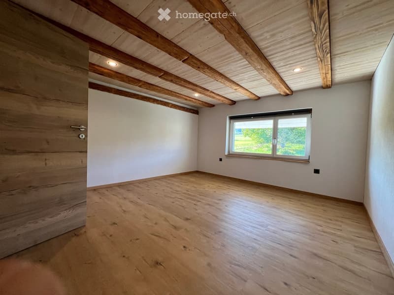 Empty room with wooden floor, exposed wooden beams on the ceiling, white walls, and a large window showing greenery outside.