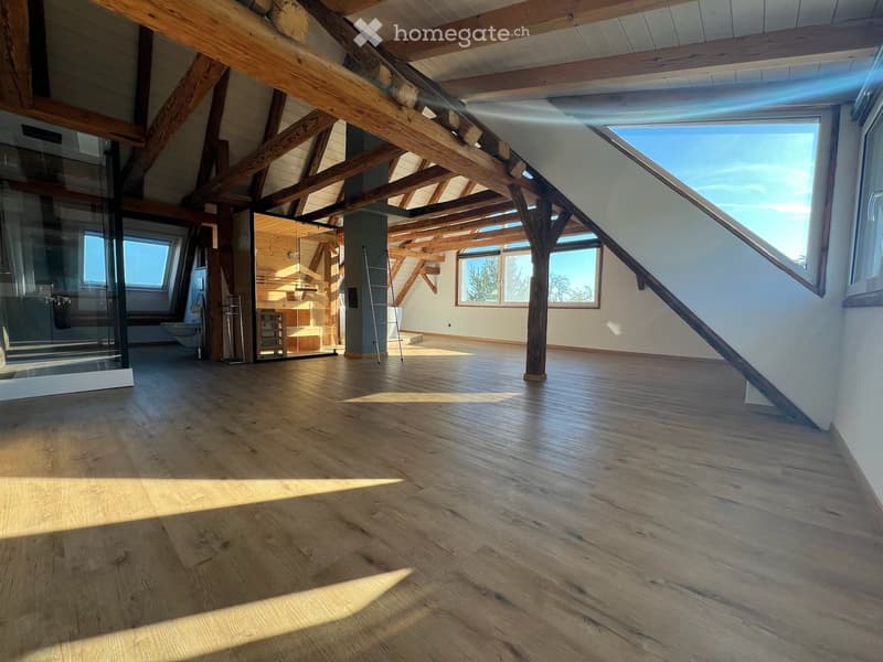 Open, airy room with exposed wooden beams and large windows, featuring a polished wooden floor and a modern glass partition leading to a bathroom area.