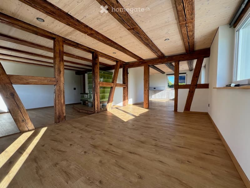 Empty room with wooden floor and exposed wooden beams on the ceiling. Natural light streams in from large windows, casting shadows on the floor.