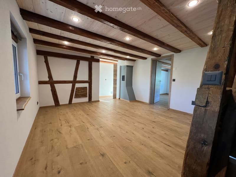 Empty room with wooden floor and exposed wooden beams on the ceiling. The room has white walls, a window on the left, and several open doorways leading to other rooms.