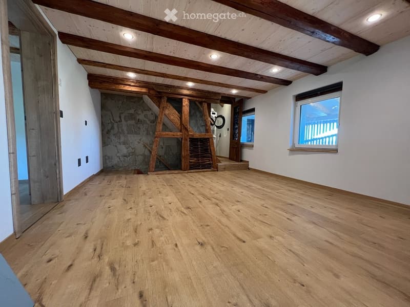 Empty room with wooden flooring, exposed wooden beams on the ceiling, white walls, and large windows allowing natural light. There is a doorway leading to another area and a small staircase visible in the background.