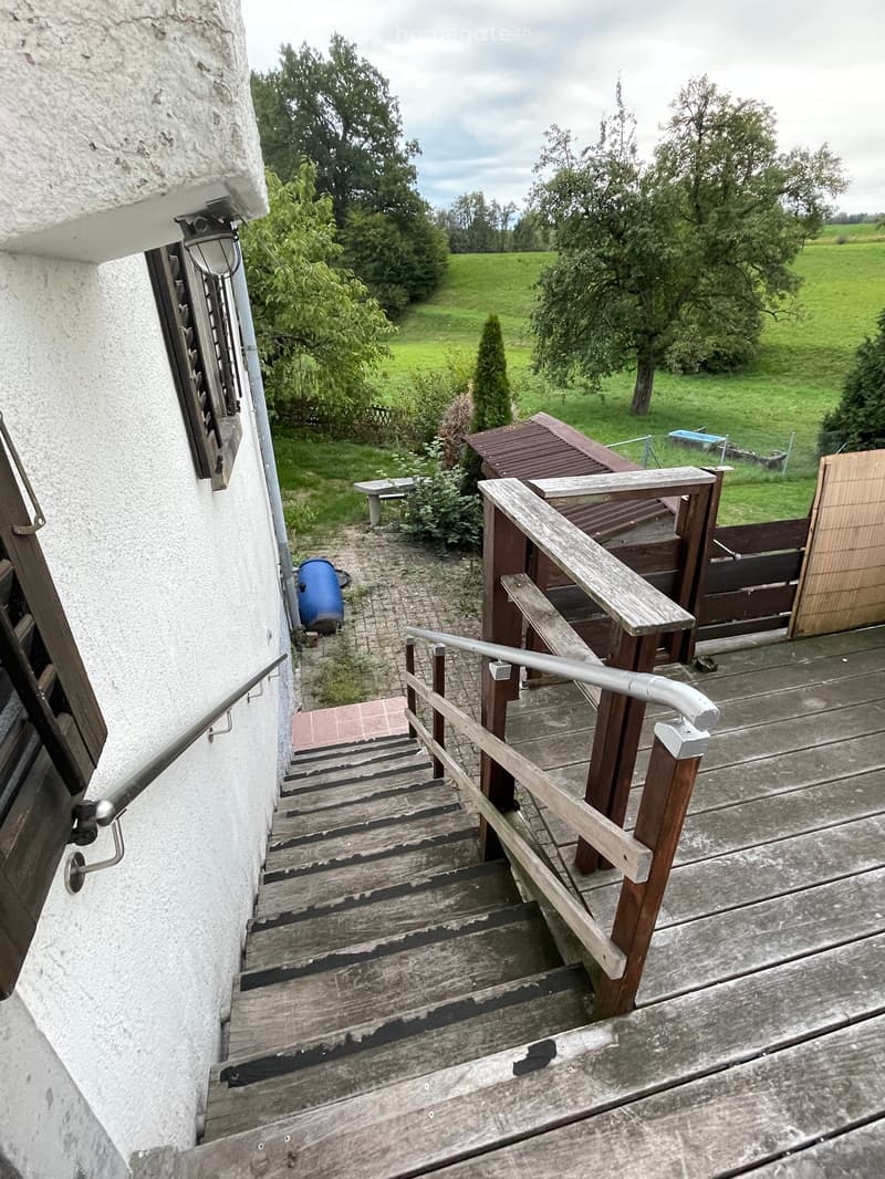 Wooden staircase leading to a wooden deck with metal railings, overlooking a grassy yard with trees and a blue barrel.