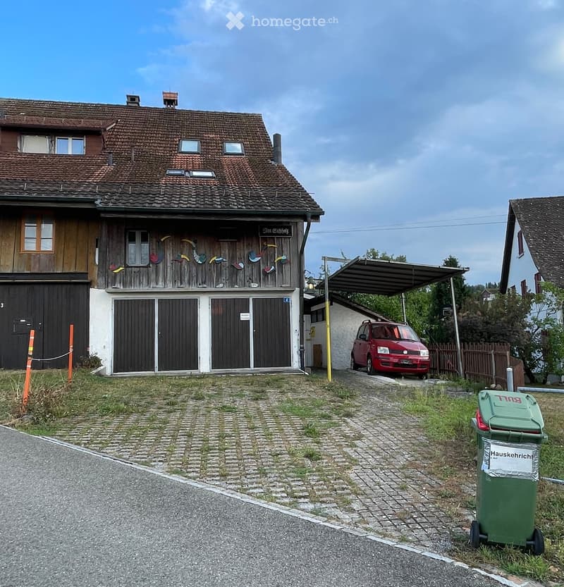 Two-story house with a tiled roof, wooden facade, and a covered parking area. A red car is parked under the carport, and there is a green trash bin nearby.