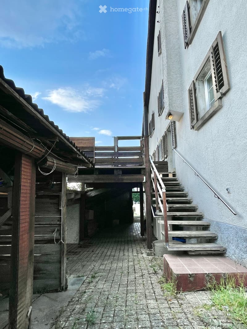 Narrow outdoor corridor with brick pavement, wooden staircase on the left, and concrete steps leading up on the right, flanked by white walls and windows.