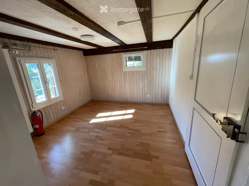 Empty room with wooden flooring, exposed wooden beams on the ceiling, and natural light from two windows. A red fire extinguisher is placed near the left wall.