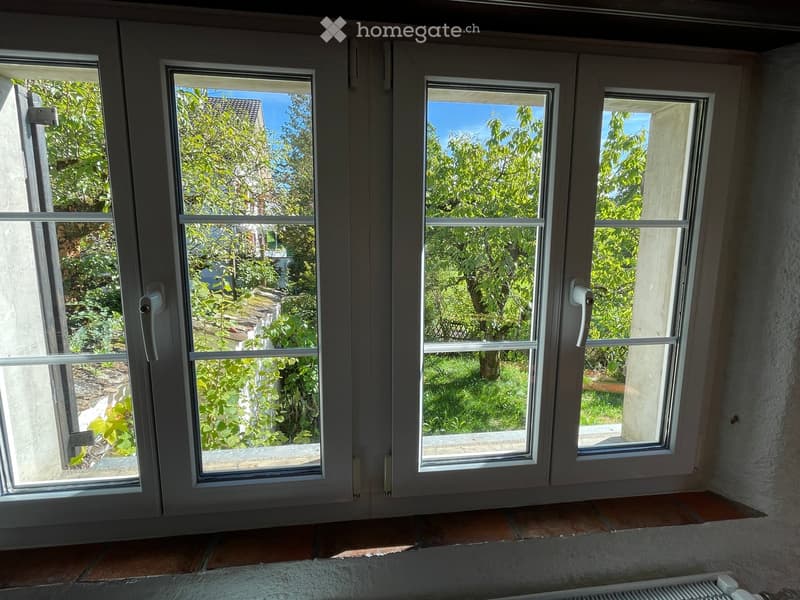 Empty room with large windows offering a view of a green, leafy outdoor area. The room has tiled flooring and a radiator visible under the windows.