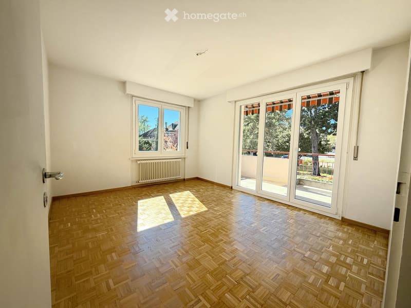 Empty room with wooden herringbone flooring, white walls, large windows, and sliding glass doors leading to a balcony.